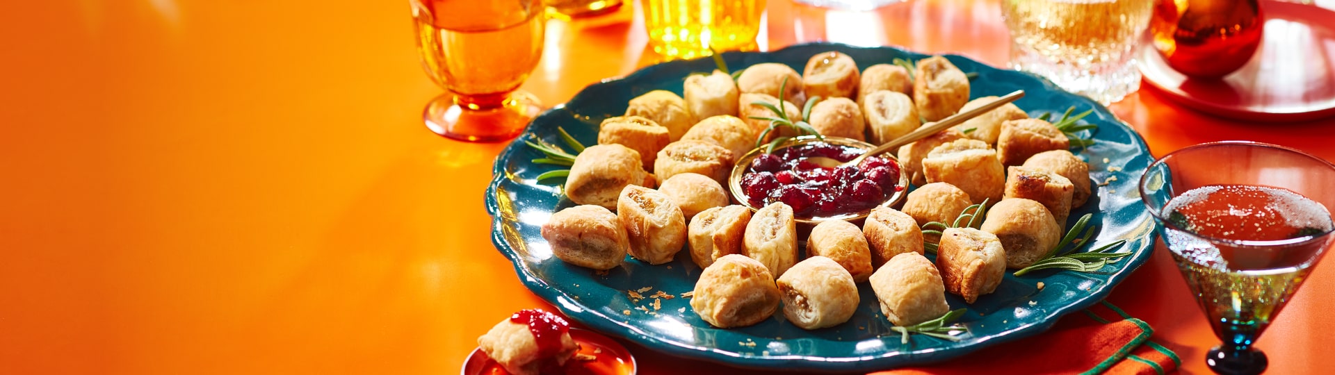 Orange surface with blue plate of sausage rolls arranged in a wreath shape with fresh rosemary and dipping sauce