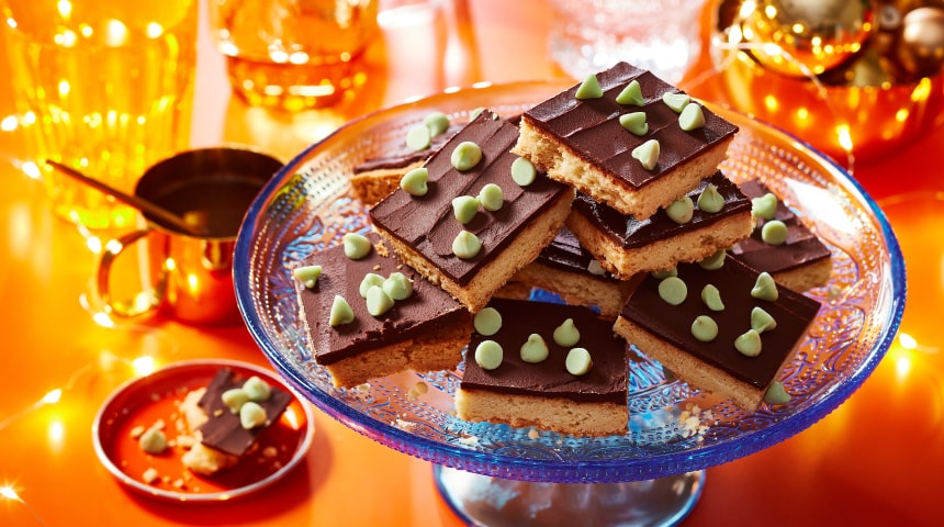 Orange table with twinkle light décor, and a blue glass cake stand topped with mint chocolate shortbread slice cookies with mint flavoured baking chips sprinkled over the top