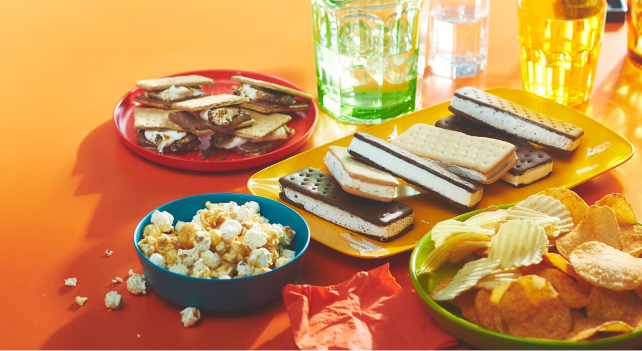 orange background with a plate of ice cream sandwiches, a bowl of chips and popcorn, and a plate of s'mores