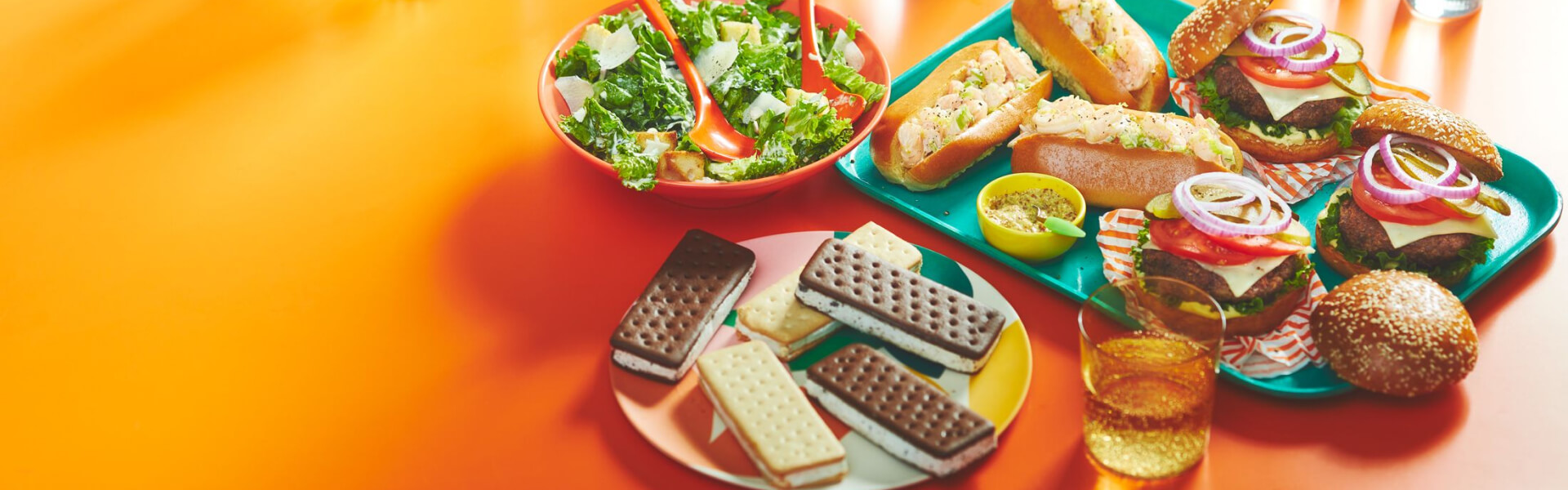 orange surface with platters of food: shrimp rolls, burgers, caesar salad and ice cream bars