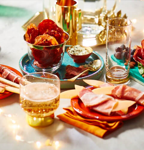 Marble table with twinkle lights, a charcuterie board, plate of salami chips, and prosciutto-wrapped melon and breadsticks.