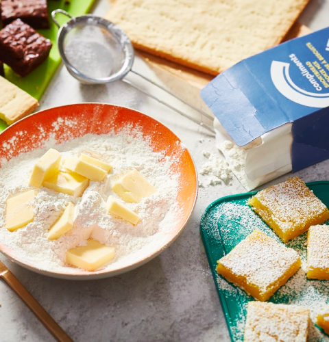 Compliments Shortbread Cookie Mix Countertop wth baking ingredients, including Compliments Shortbread Cookie Mix, a bowl with cubed butter and a knife.