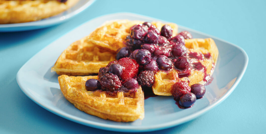 Blue tabletop with a blue breakfast plate with a cooked waffle topped in mixed berry fruit sauce.