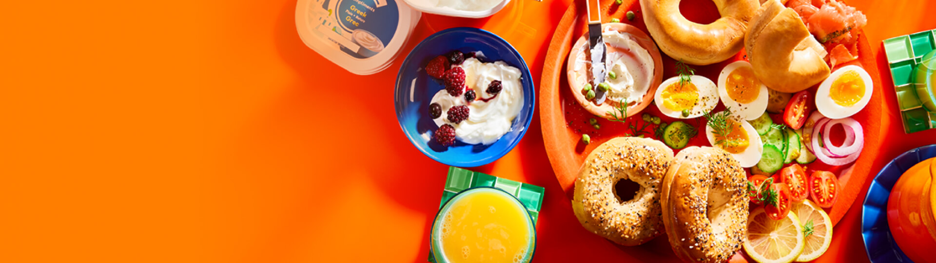 Orange background with breakfast setting including juice, tea, a cream cheese-spread bagel with smoked salmon, dill and capers.