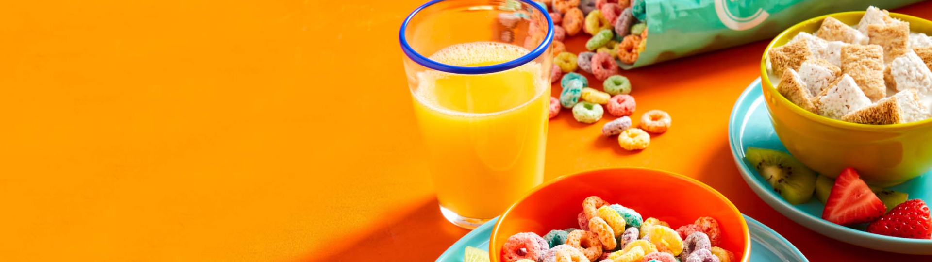 orange background with open bag of Compliments fruity hoops cereal, two bowls of cereal with milk, and a glass of orange juice