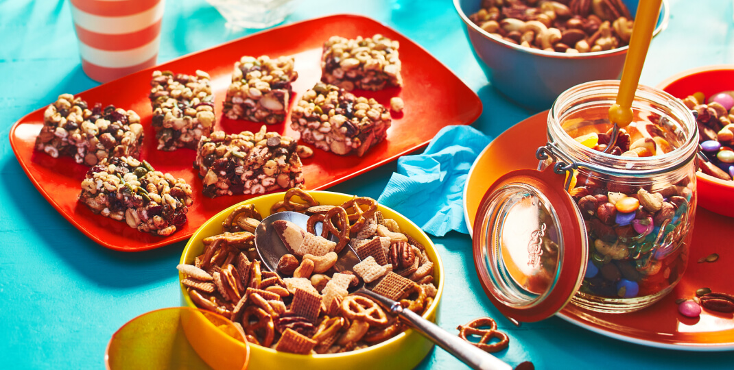 aqua blue tabletop with a plate of mixed nut snack bars, and two bowls of trail mix
