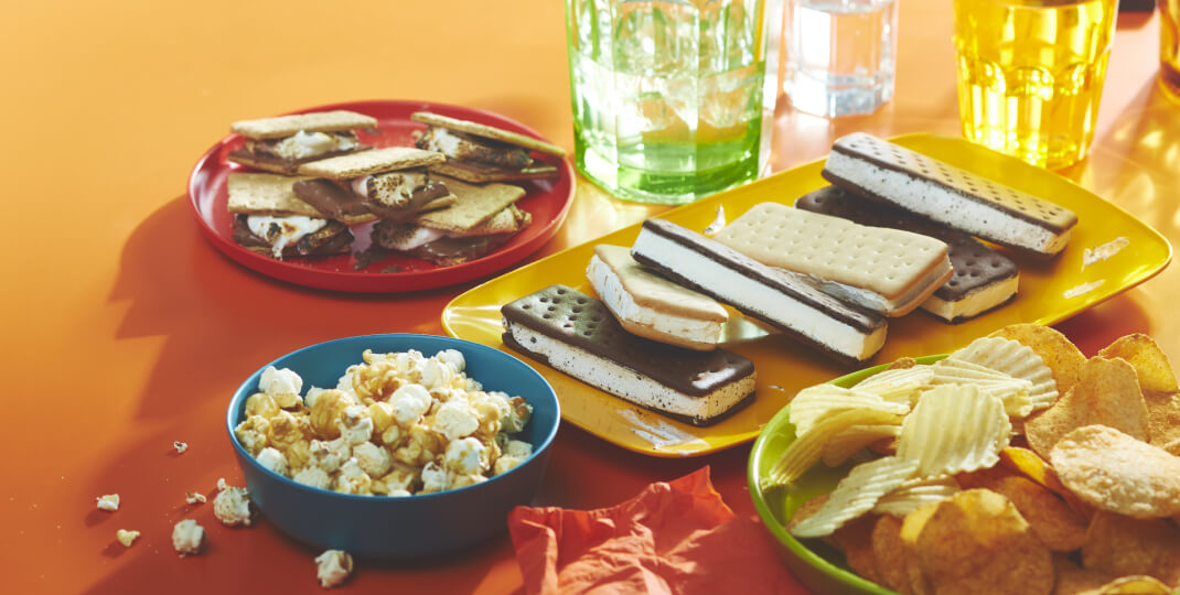 orange background with a plate of ice cream sandwiches, a bowl of chips and popcorn, and a plate of s'mores