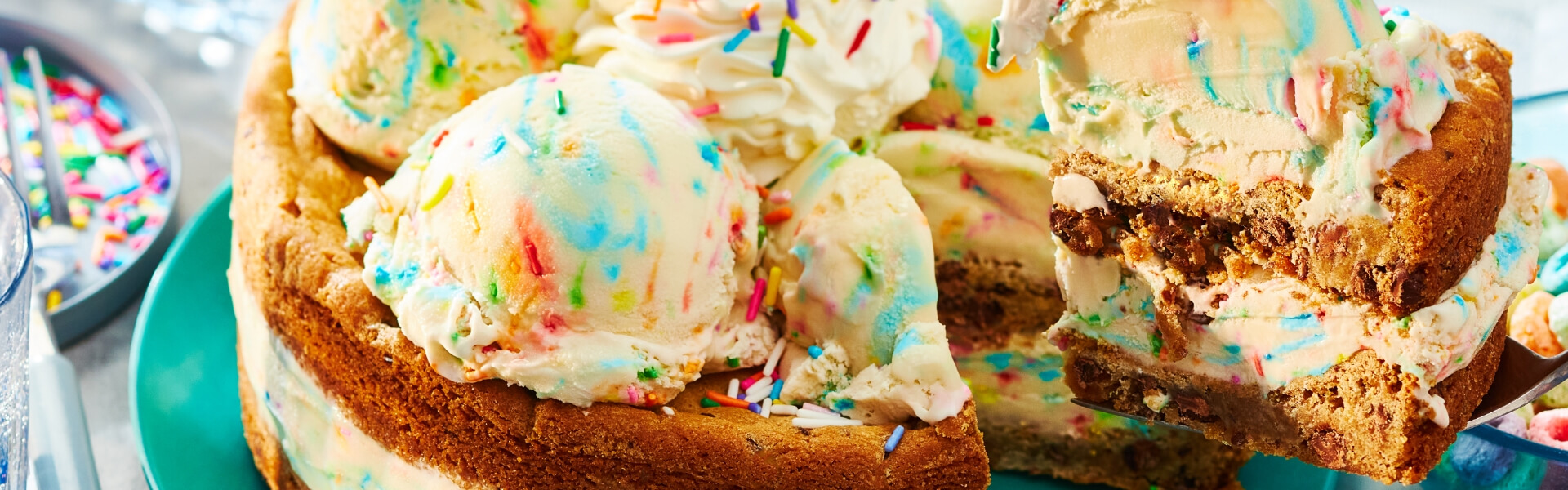 a hand holding an ice cream scoop over a tub of Compliments birthday cake flavour ice cream, pulling it through to scoop out white and coloured sprinkle scoop of ice cream, with cones ona plate in the background