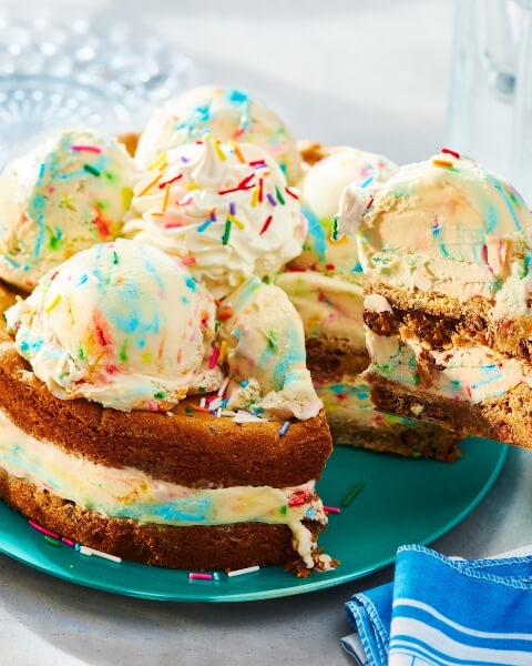 Summer_try__me a hand holding an ice cream scoop over a tub of Compliments birthday cake flavour ice cream, pulling it through to scoop out white and coloured sprinkle scoop of ice cream, with cones ona plate in the background