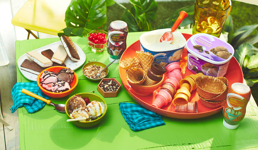 build an epic sundae station Green outdoor table with an array of ice cream sundae ingredients including ice cream, sundae sauces, cones, and toppings